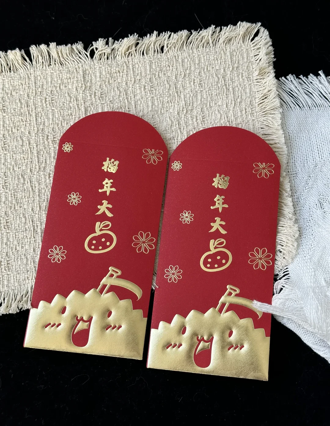 两只并排的“榴年大吉”红包实拍:红色纸面上是烫金与浮雕图案,放在米白色织物上